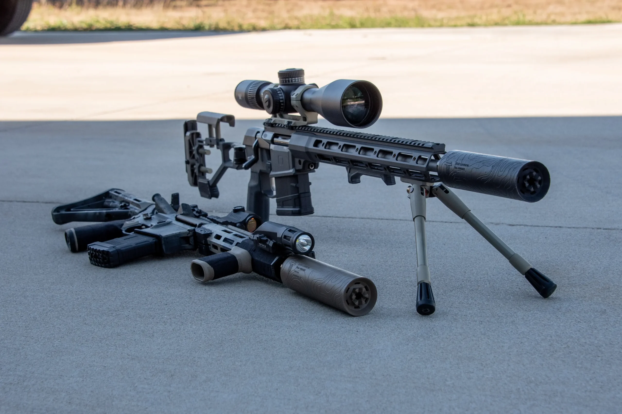 A suppressed bolt rifle on a bipod and a suppressed AR beside it on a tarmac